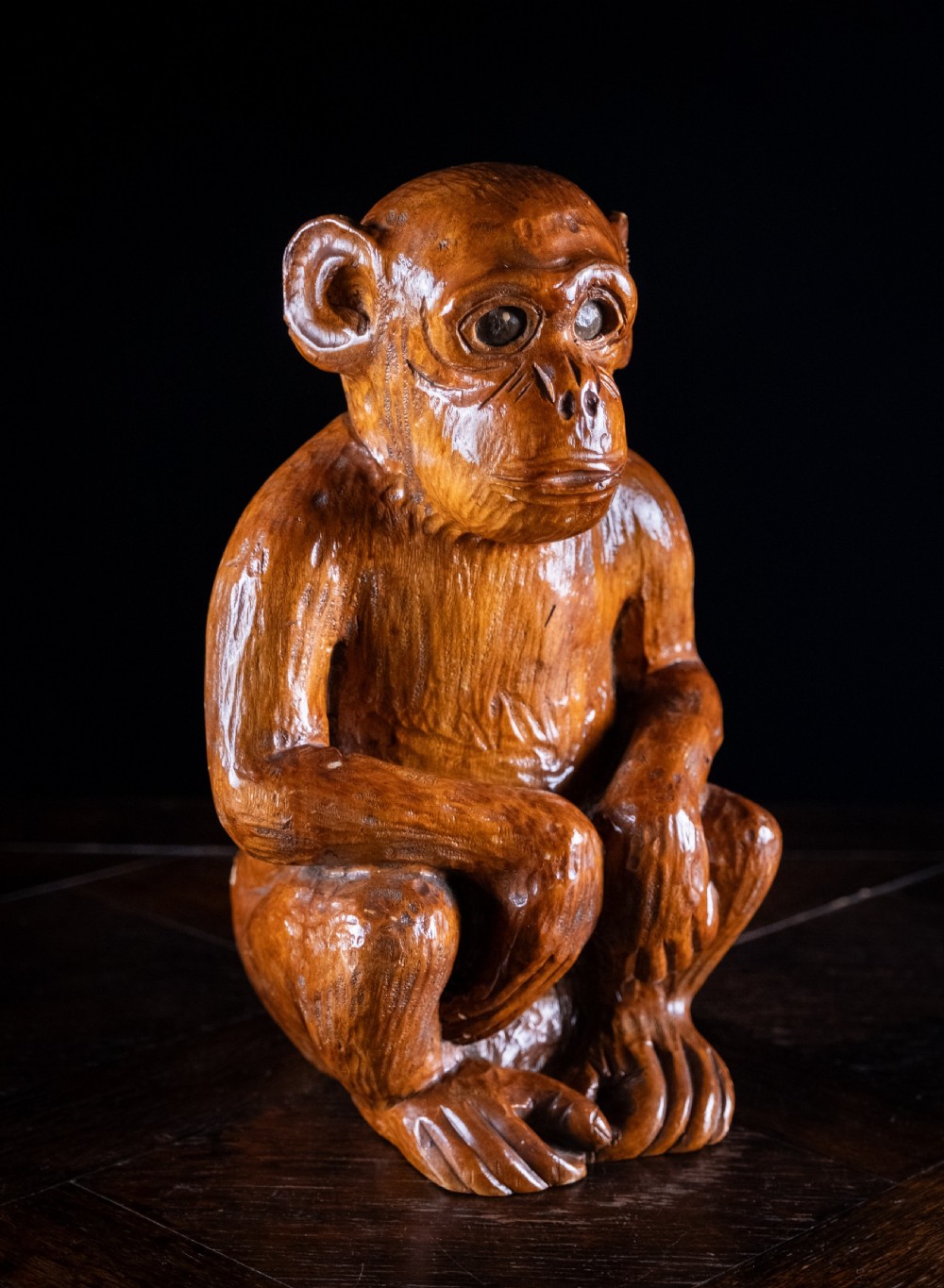 A hand-carved warm brown wooden sculpture depicting a seated monkey with chiselled fur details and dark eyes. View IX
