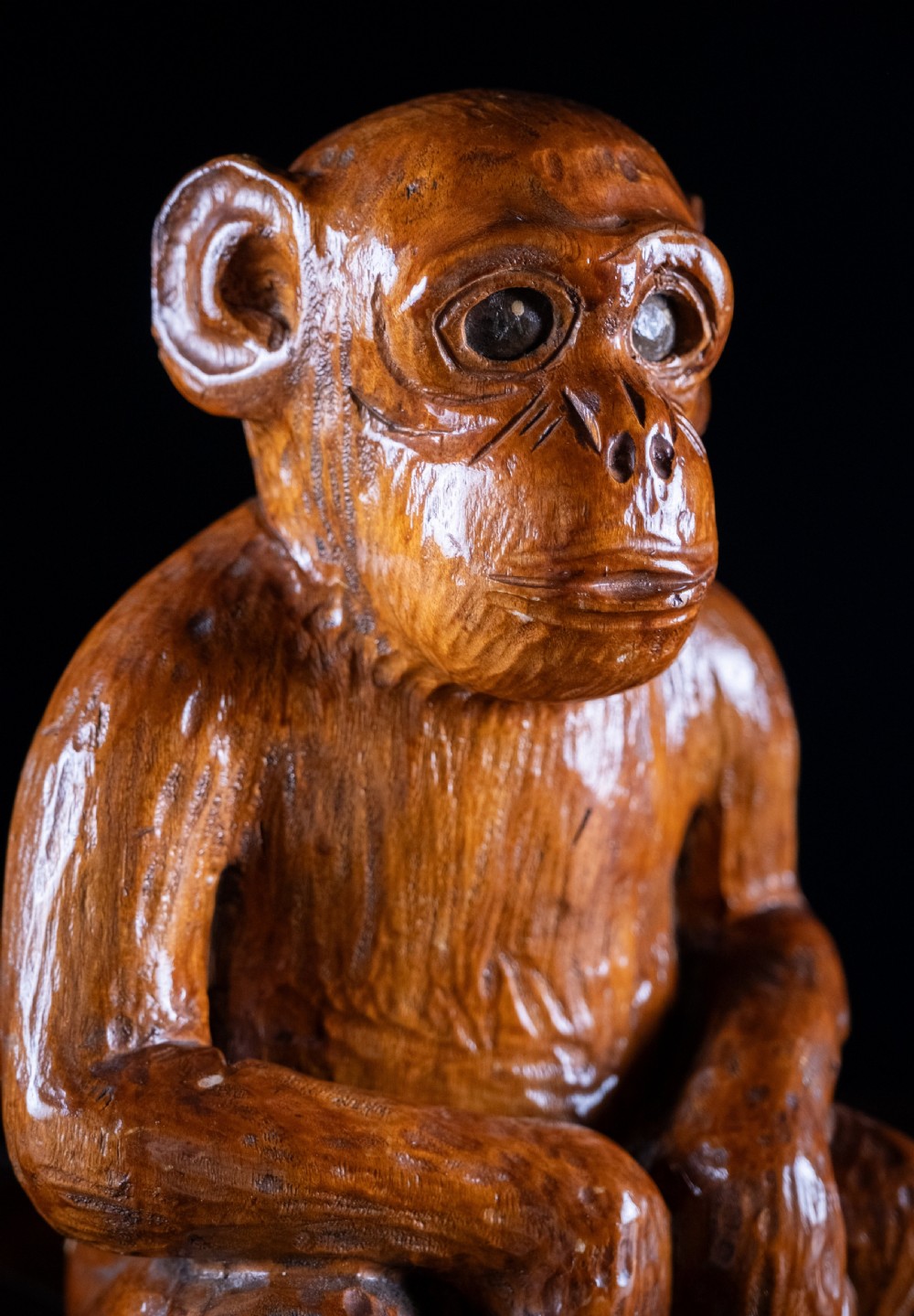 A hand-carved warm brown wooden sculpture depicting a seated monkey with chiselled fur details and dark eyes. View X