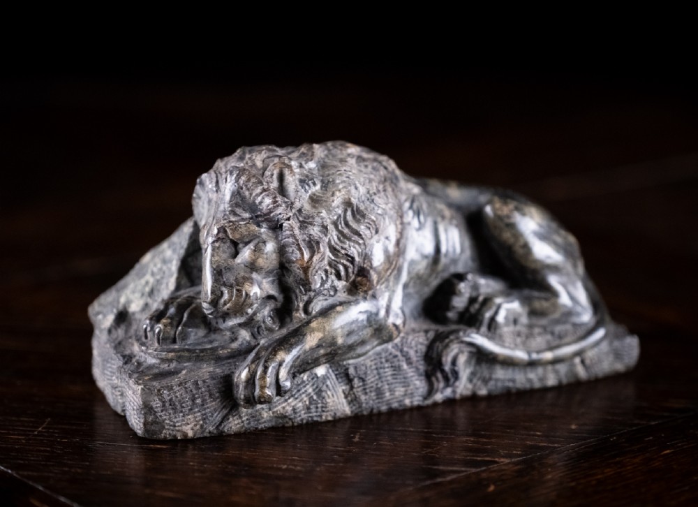 A carved dark serpentine stone sculpture depicting the recumbent Lion of Lucerne resting on an integral rectangular stone base. View III