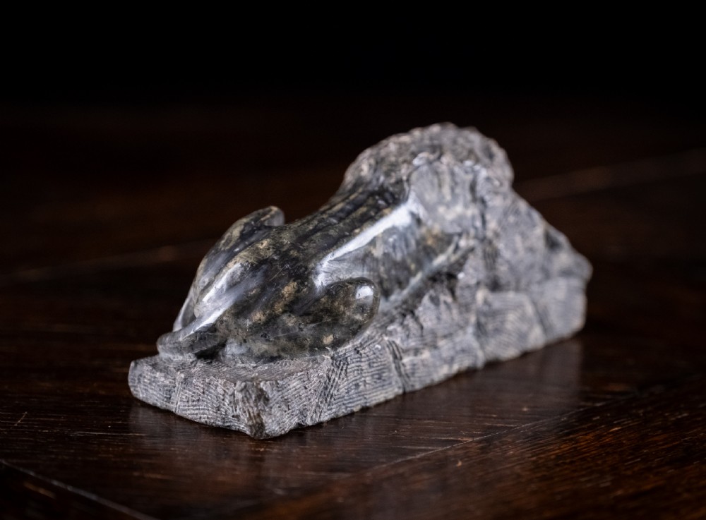 A carved dark serpentine stone sculpture depicting the recumbent Lion of Lucerne resting on an integral rectangular stone base. View IV