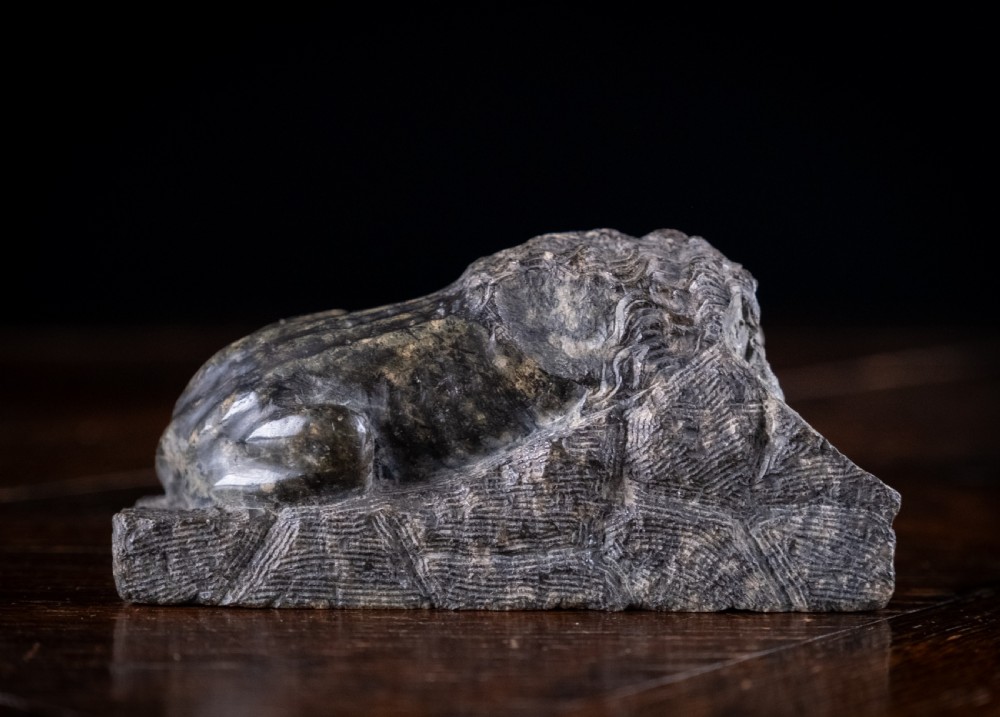 A carved dark serpentine stone sculpture depicting the recumbent Lion of Lucerne resting on an integral rectangular stone base. View VI