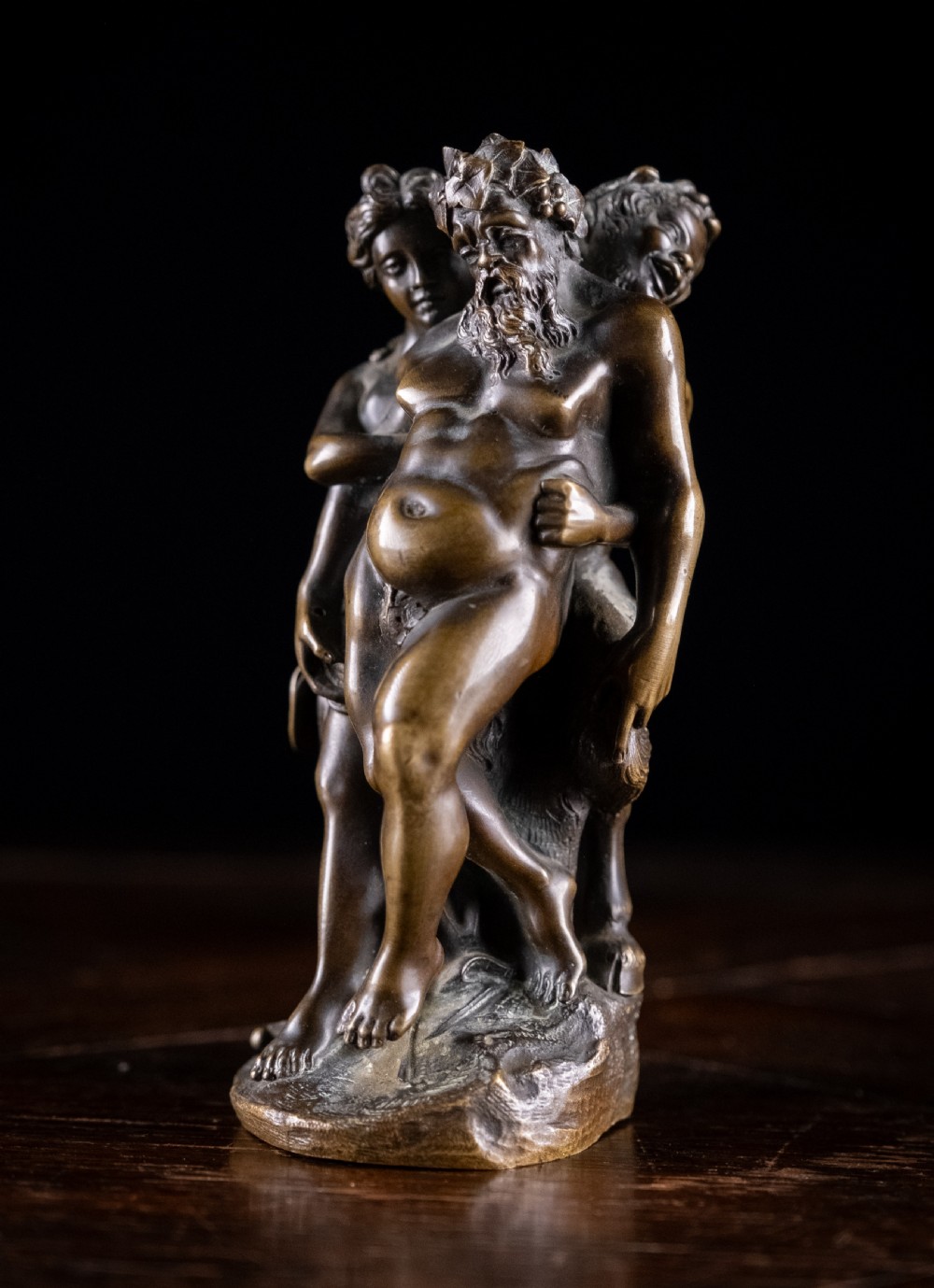 A dark brown bronze figurative sculpture group depicting Silenus supported by Pan and a Nereid on a textured bronze base.