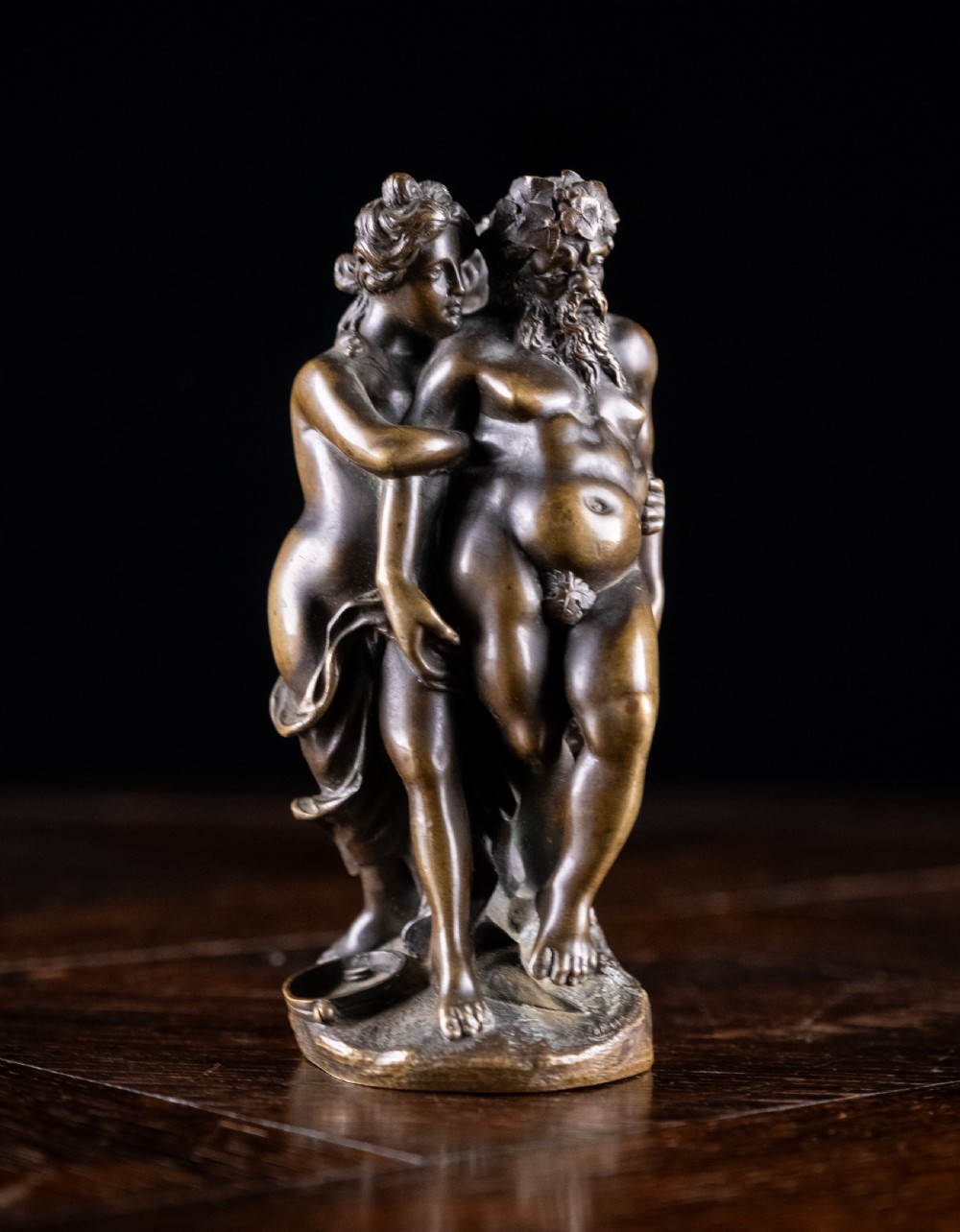A dark brown bronze figurative sculpture group depicting Silenus supported by Pan and a Nereid on a textured bronze base. View II