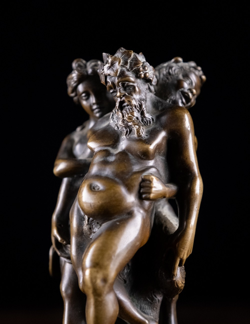A dark brown bronze figurative sculpture group depicting Silenus supported by Pan and a Nereid on a textured bronze base. View IV