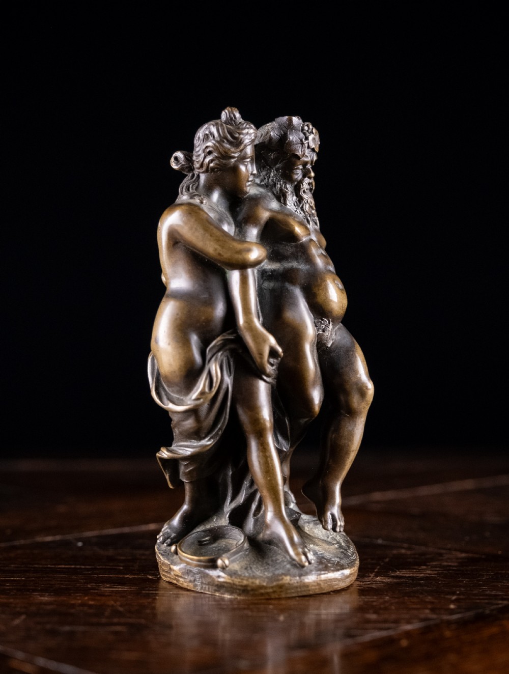 A dark brown bronze figurative sculpture group depicting Silenus supported by Pan and a Nereid on a textured bronze base. View X