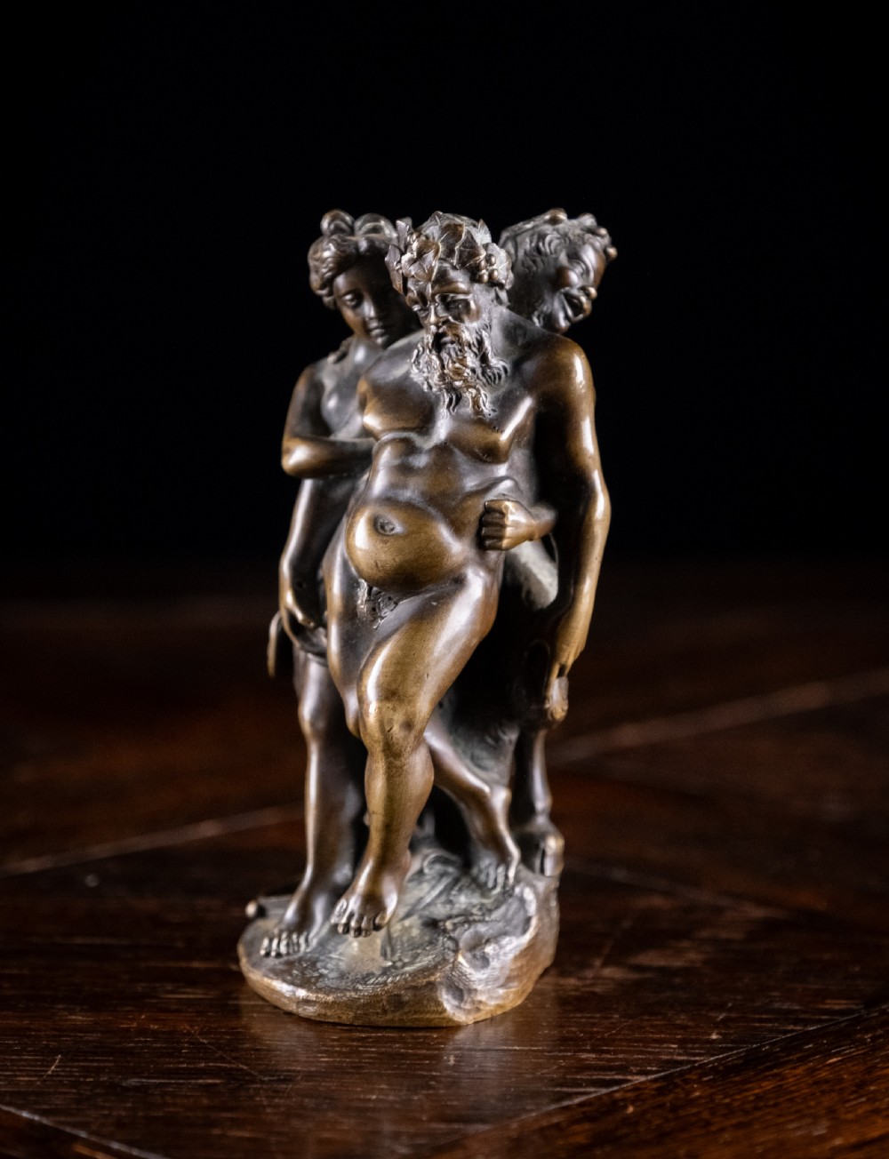 A dark brown bronze figurative sculpture group depicting Silenus supported by Pan and a Nereid on a textured bronze base. View XI