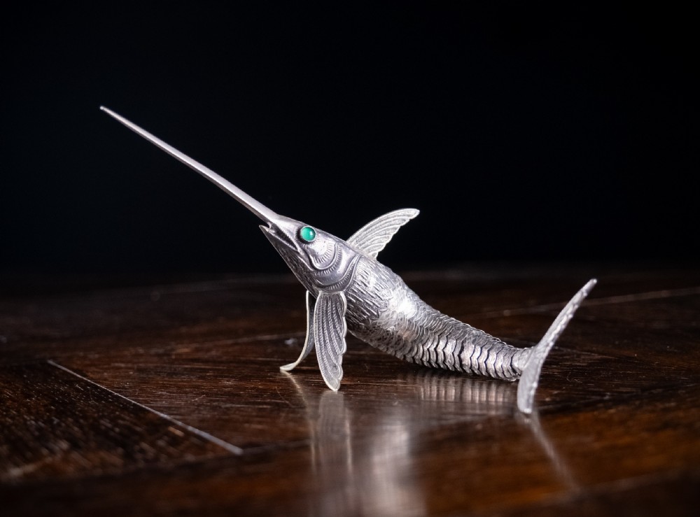 An articulated silver swordfish figure with detailed scales, fins, extended bill, and green stone eyes resting on a dark surface.