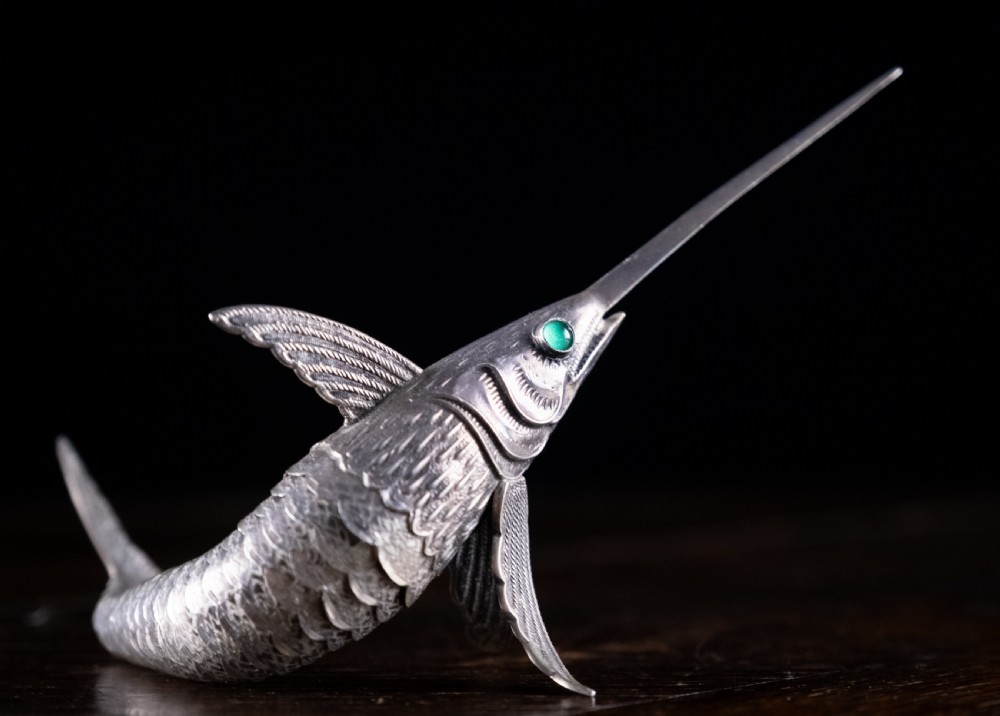 An articulated silver swordfish figure with detailed scales, fins, extended bill, and green stone eyes resting on a dark surface. View II