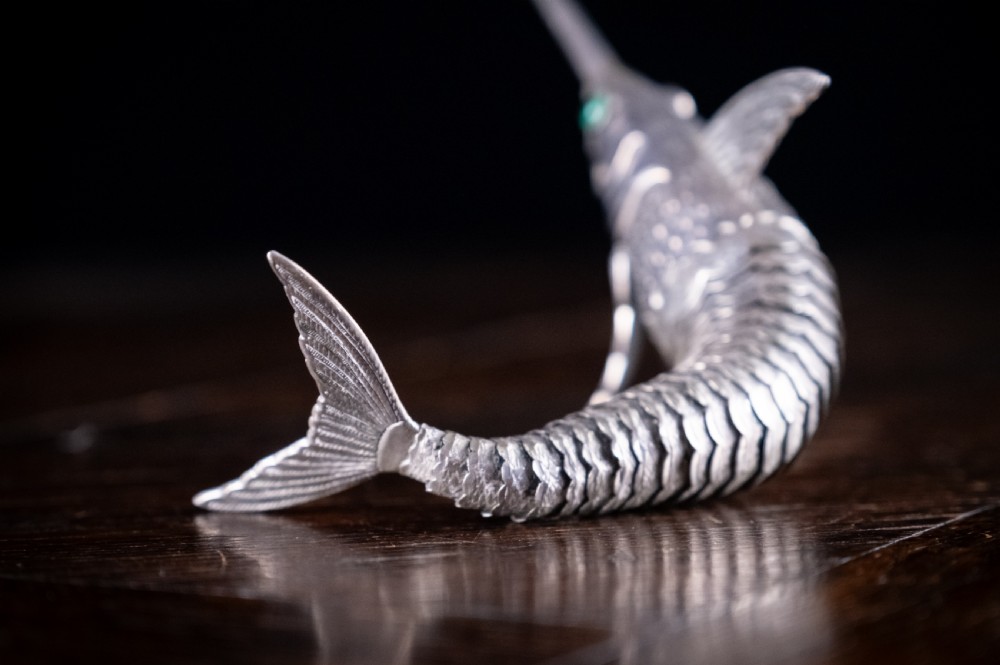 An articulated silver swordfish figure with detailed scales, fins, extended bill, and green stone eyes resting on a dark surface. View III