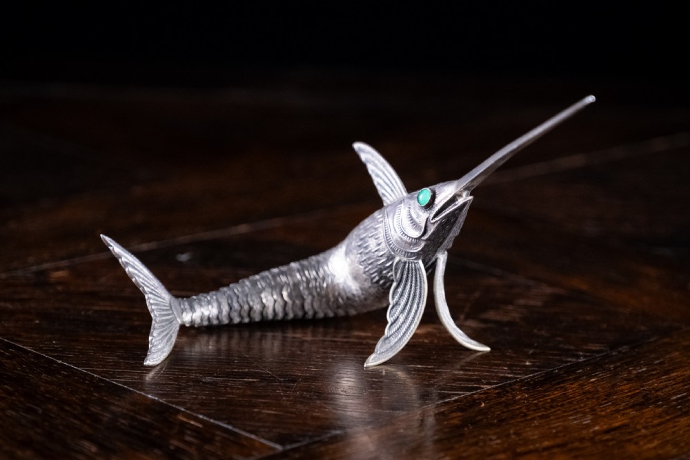 An articulated silver swordfish figure with detailed scales, fins, extended bill, and green stone eyes resting on a dark surface. View IV