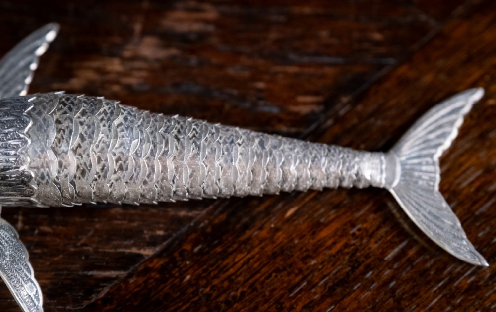 An articulated silver swordfish figure with detailed scales, fins, extended bill, and green stone eyes resting on a dark surface. View VII