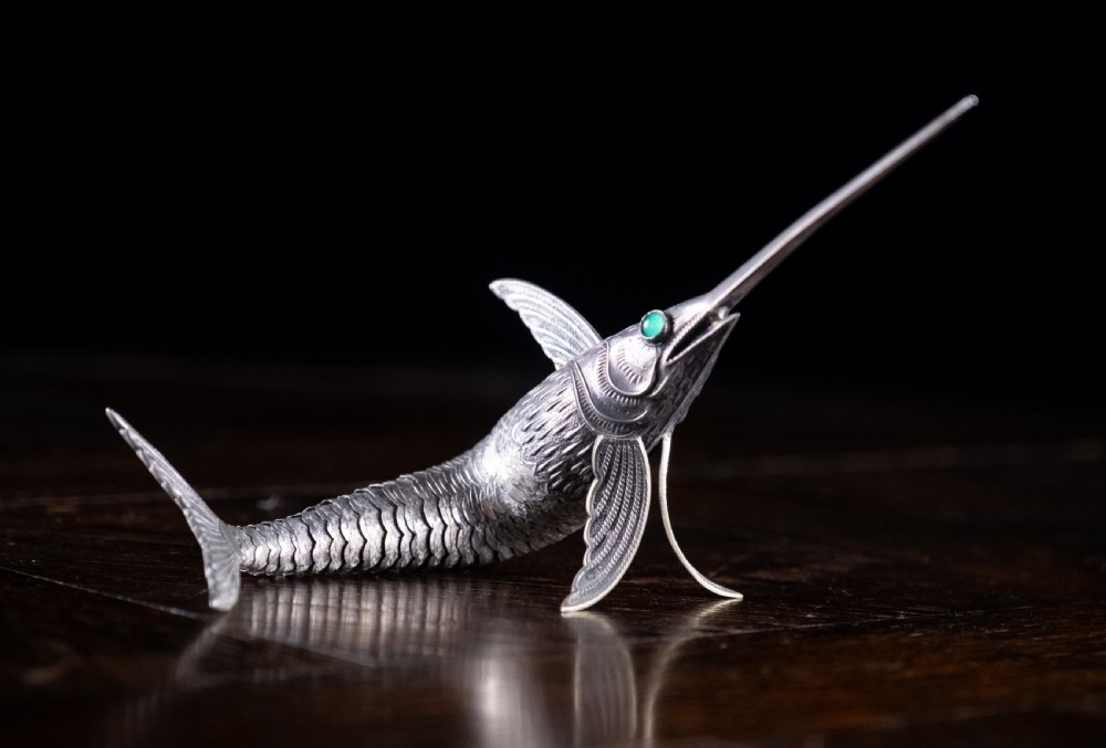 An articulated silver swordfish figure with detailed scales, fins, extended bill, and green stone eyes resting on a dark surface. View VIII