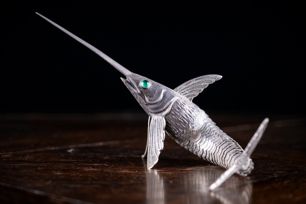 An articulated silver swordfish figure with detailed scales, fins, extended bill, and green stone eyes resting on a dark surface. View IX