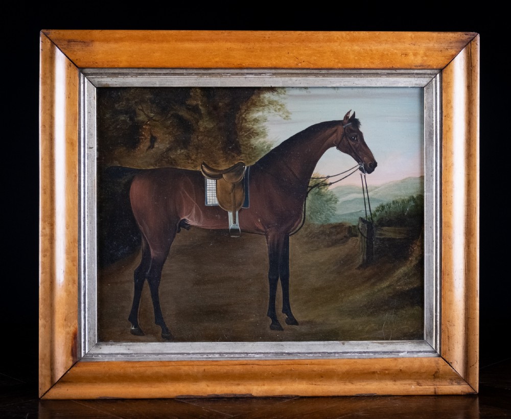 Early 19th-century oil painting on board depicting a saddled bay hunter horse standing in profile within a rural landscape, presented in a light wood frame.