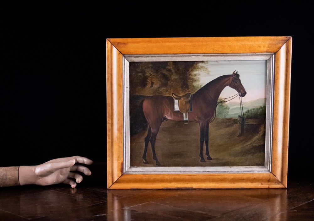 Early 19th-century oil painting on board depicting a saddled bay hunter horse standing in profile within a rural landscape, presented in a light wood frame. View IV