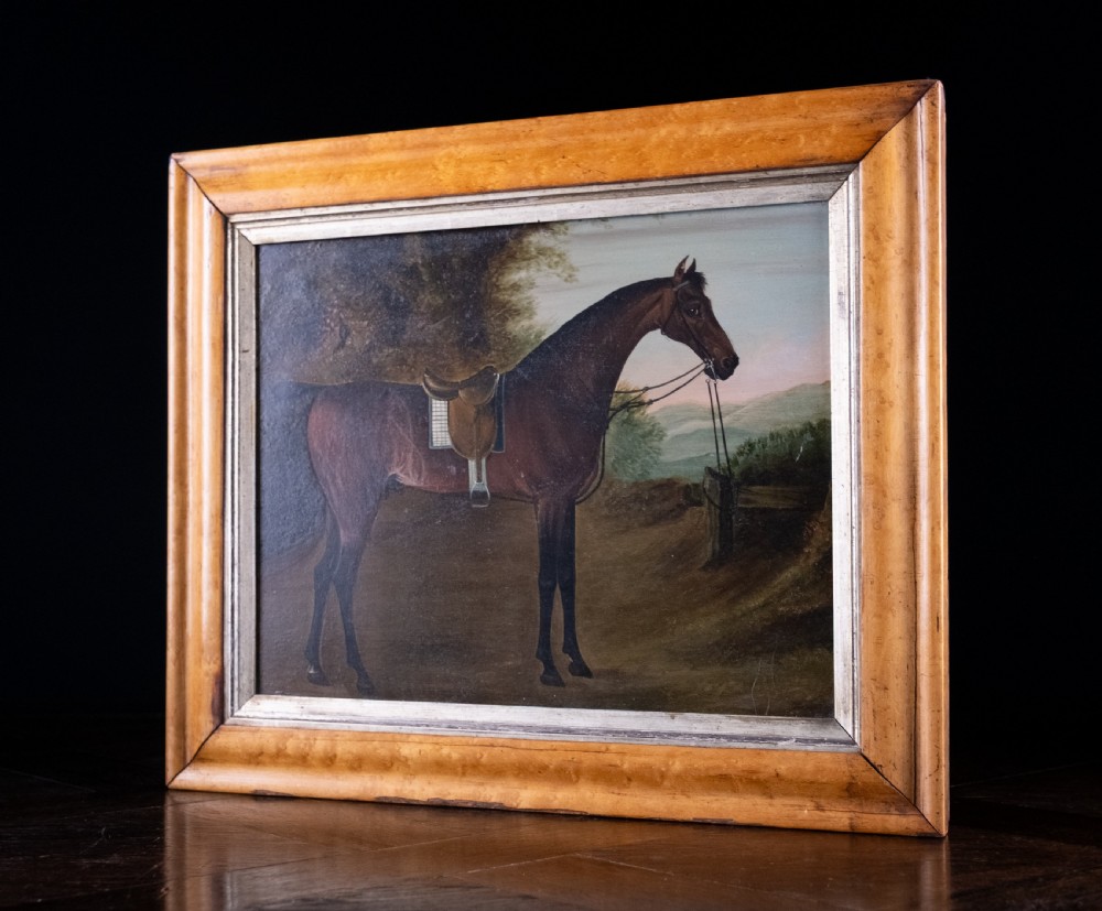 Early 19th-century oil painting on board depicting a saddled bay hunter horse standing in profile within a rural landscape, presented in a light wood frame. View VI