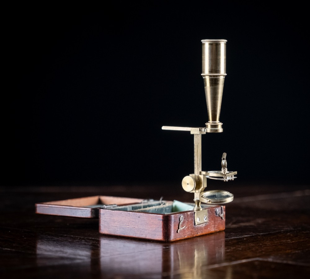 A small early 19th-century lacquered brass pocket microscope mounted onto the rim of its fitted mahogany storage box.