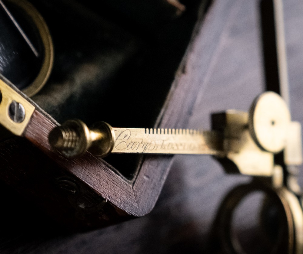 A small early 19th-century lacquered brass pocket microscope mounted onto the rim of its fitted mahogany storage box. View V