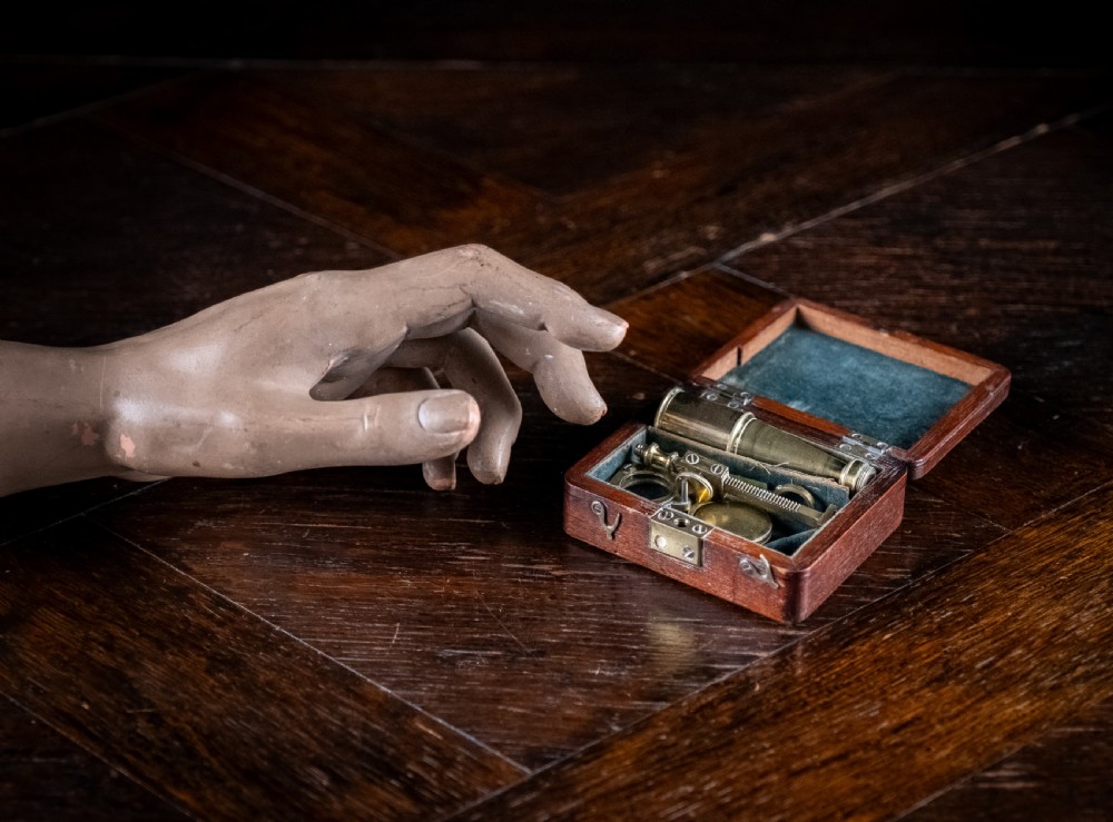 A small early 19th-century lacquered brass pocket microscope mounted onto the rim of its fitted mahogany storage box. View X