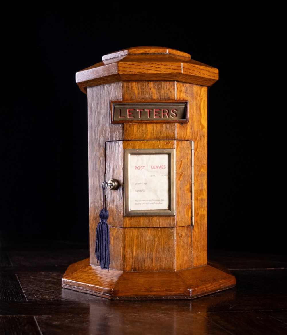 An octagonal oak tabletop post box featuring a brass letter slot marked LETTERS and a key-locked front door with a collection schedule frame. View III
