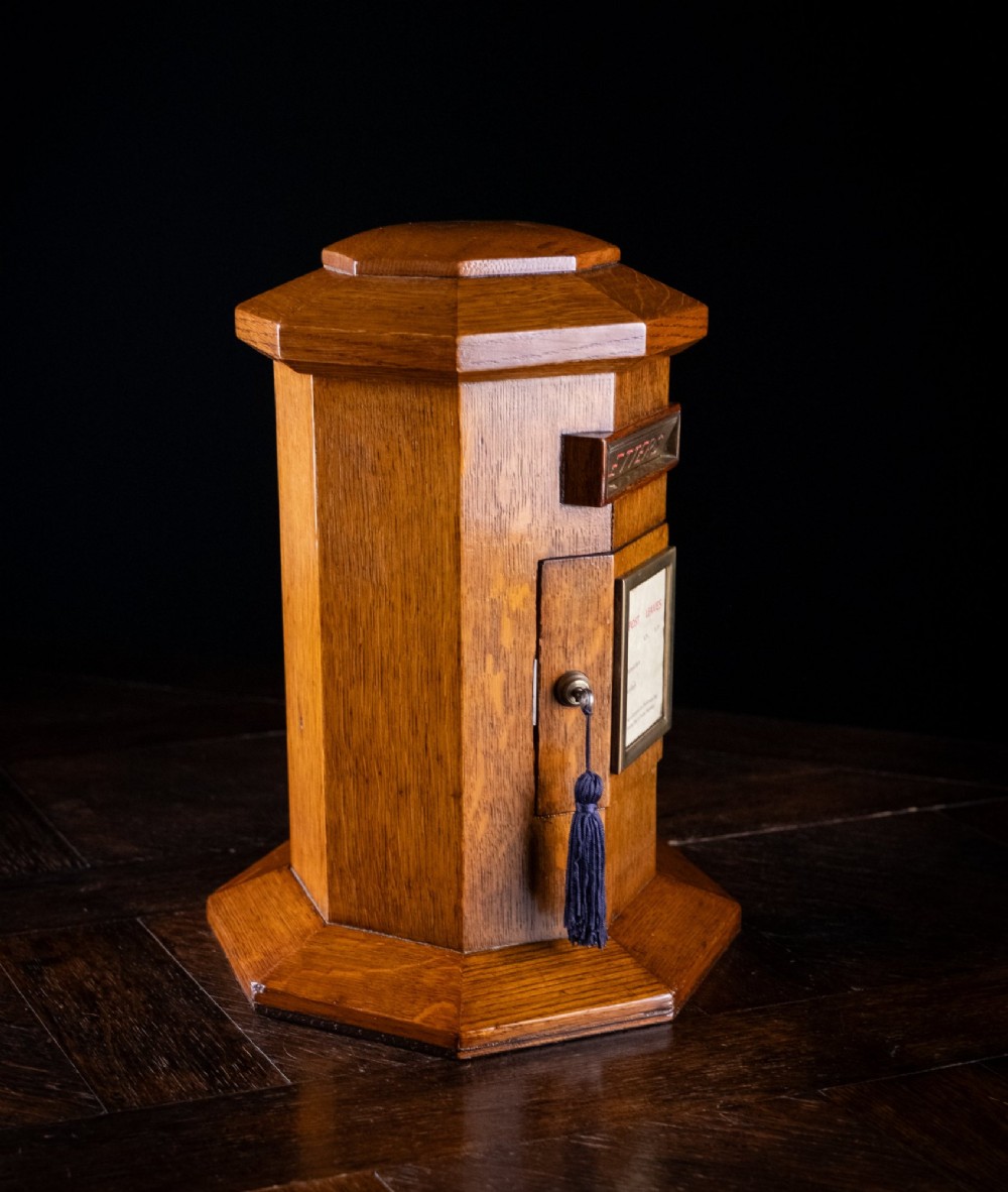 An octagonal oak tabletop post box featuring a brass letter slot marked LETTERS and a key-locked front door with a collection schedule frame. View IV