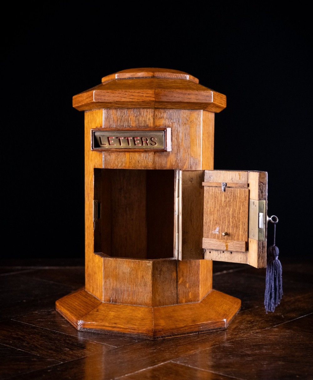 An octagonal oak tabletop post box featuring a brass letter slot marked LETTERS and a key-locked front door with a collection schedule frame. View VII