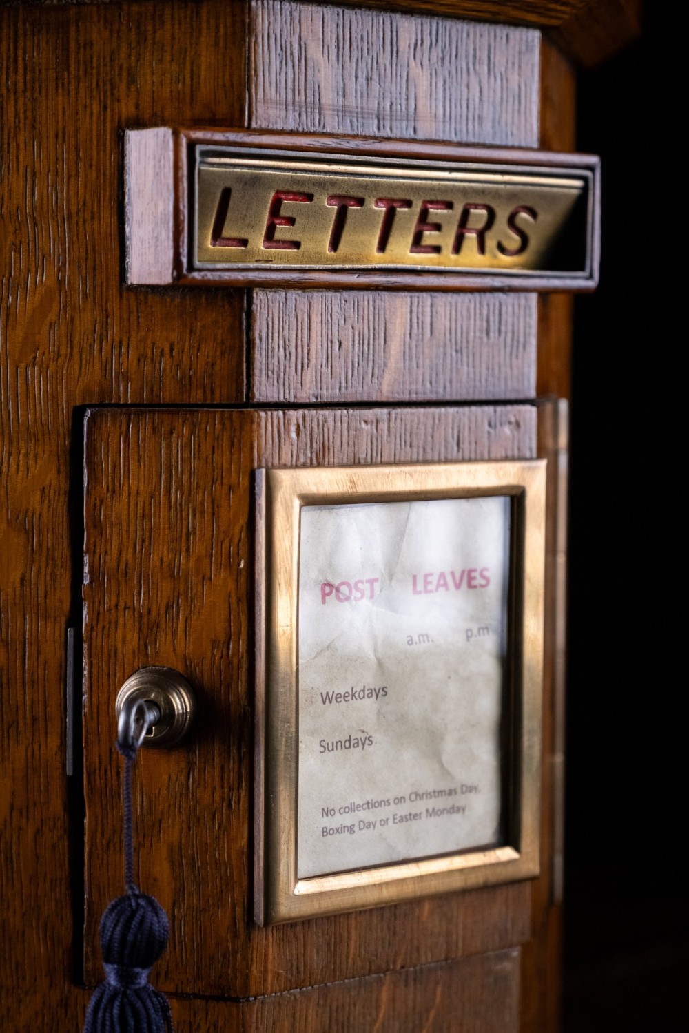 An octagonal oak tabletop post box featuring a brass letter slot marked LETTERS and a key-locked front door with a collection schedule frame. View VIII