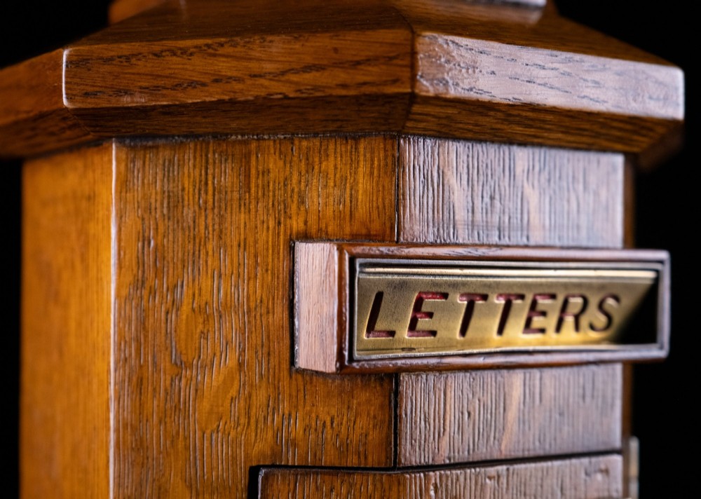 An octagonal oak tabletop post box featuring a brass letter slot marked LETTERS and a key-locked front door with a collection schedule frame. View IX