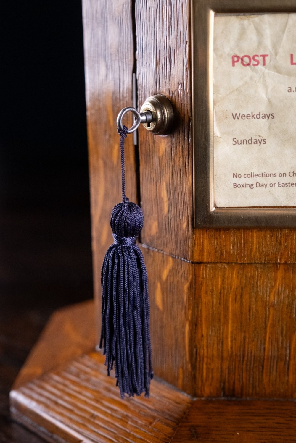 An octagonal oak tabletop post box featuring a brass letter slot marked LETTERS and a key-locked front door with a collection schedule frame. View X