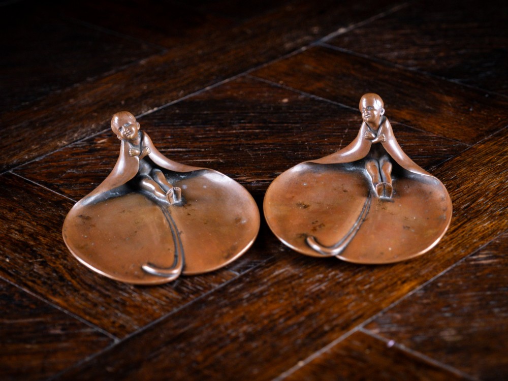 A pair of small, patinated bronze leaf-shaped dishes, each featuring a sculpted figure of a child seated at the folded edge.