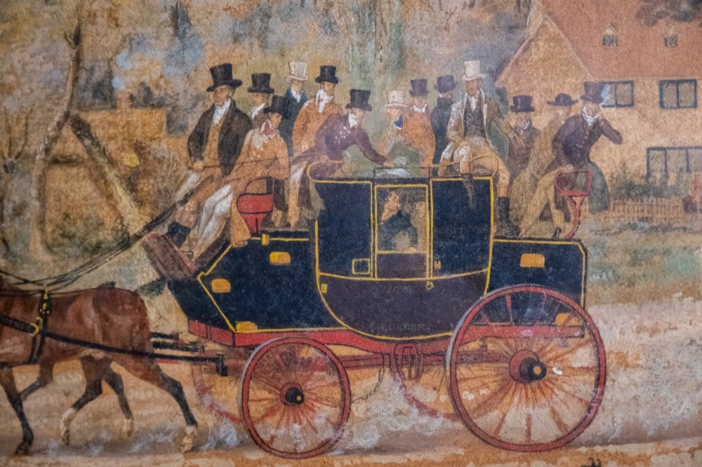 A framed 19th-century oil painting on board depicting a horse-drawn stagecoach travelling along a rural dirt road past figures and a cottage under a cloudy sky. View III