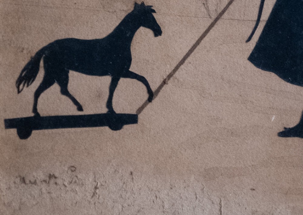A 19th-century cut-paper silhouette depicting a young child pulling a toy horse on wheels, set within a bevelled wooden frame with a gilded inner slip. View III
