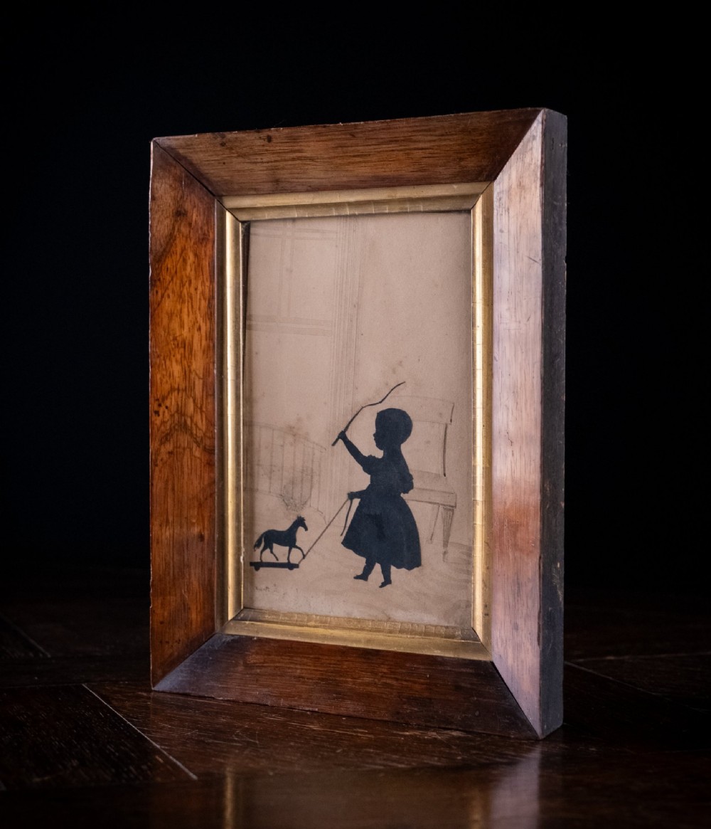 A 19th-century cut-paper silhouette depicting a young child pulling a toy horse on wheels, set within a bevelled wooden frame with a gilded inner slip. View IV