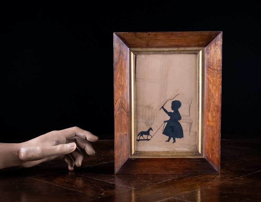 A 19th-century cut-paper silhouette depicting a young child pulling a toy horse on wheels, set within a bevelled wooden frame with a gilded inner slip. View V