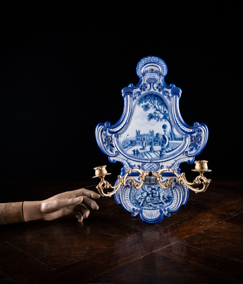 A Rococo Revival blue and white glazed ceramic wall plaque depicting an architectural scene and cherubs, fitted with two gilt ormolu candle branches. View II