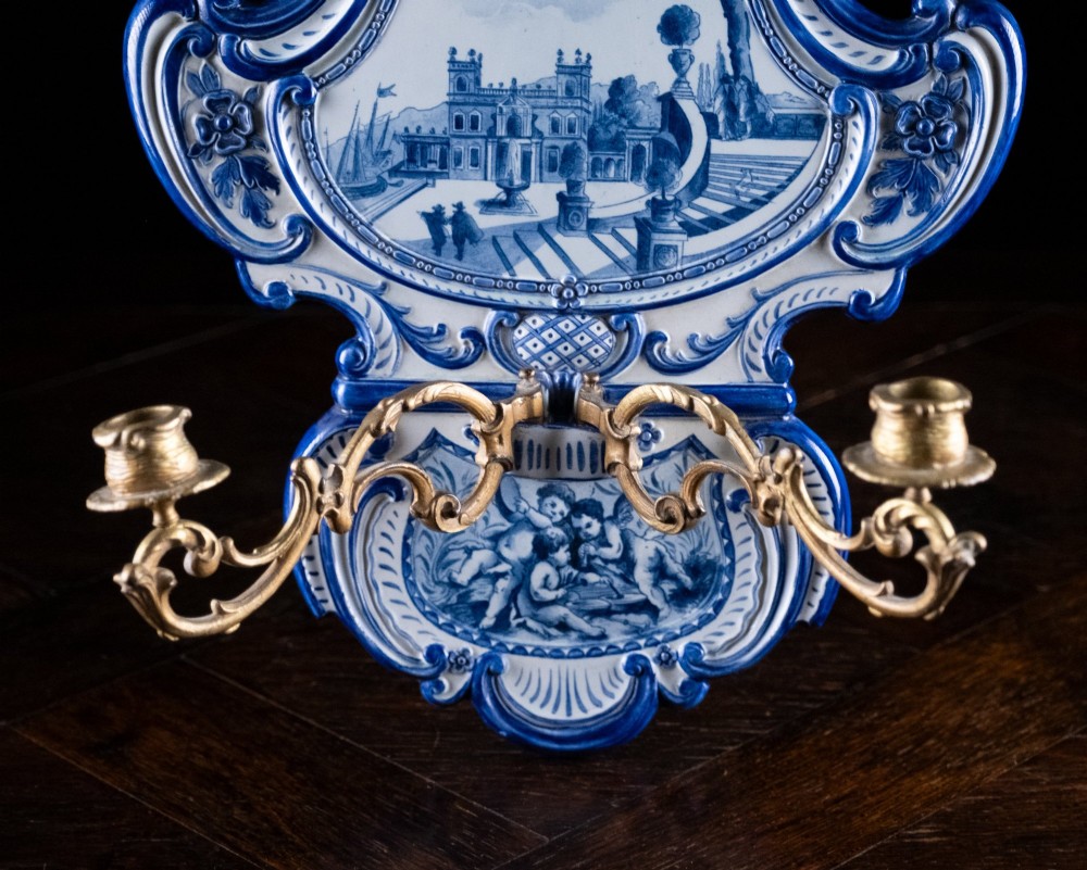 A Rococo Revival blue and white glazed ceramic wall plaque depicting an architectural scene and cherubs, fitted with two gilt ormolu candle branches. View V