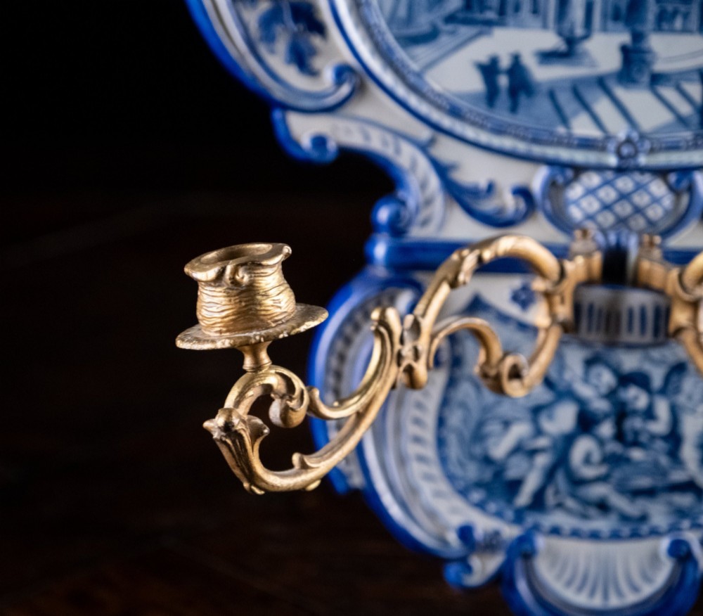 A Rococo Revival blue and white glazed ceramic wall plaque depicting an architectural scene and cherubs, fitted with two gilt ormolu candle branches. View VI