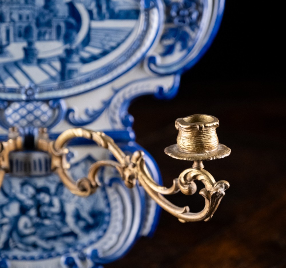 A Rococo Revival blue and white glazed ceramic wall plaque depicting an architectural scene and cherubs, fitted with two gilt ormolu candle branches. View VII
