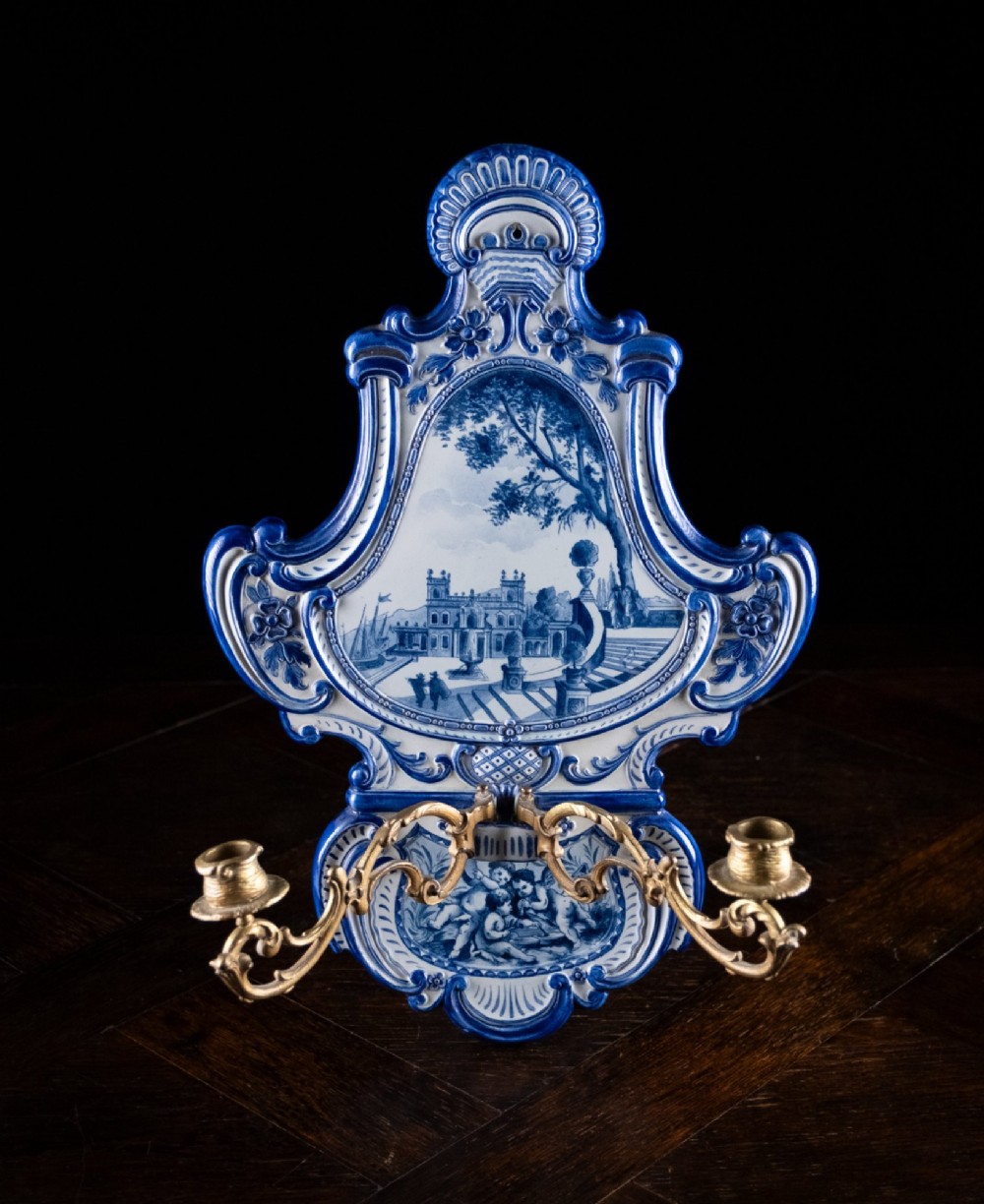 A Rococo Revival blue and white glazed ceramic wall plaque depicting an architectural scene and cherubs, fitted with two gilt ormolu candle branches. View VIII
