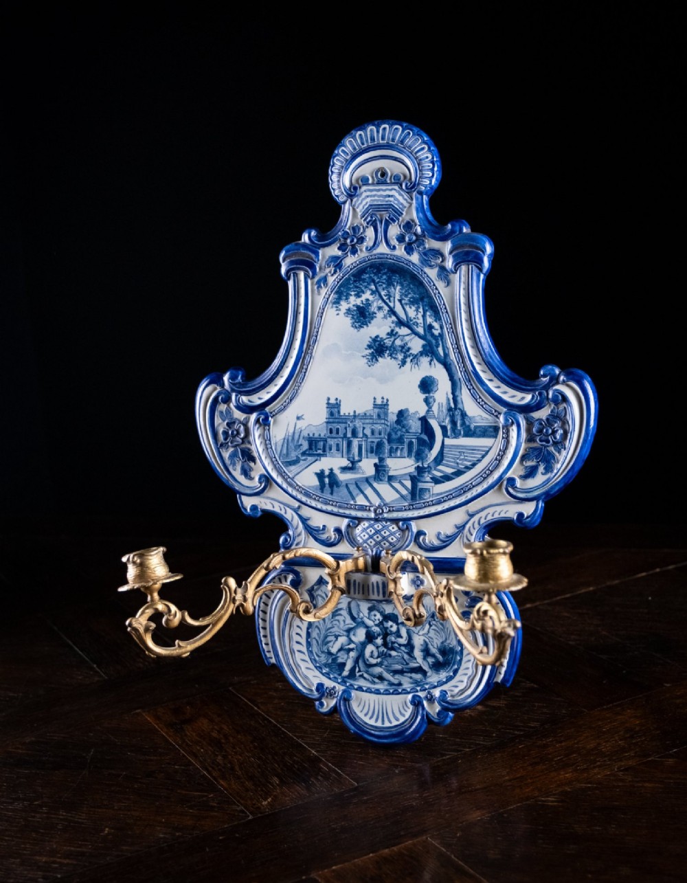 A Rococo Revival blue and white glazed ceramic wall plaque depicting an architectural scene and cherubs, fitted with two gilt ormolu candle branches. View X