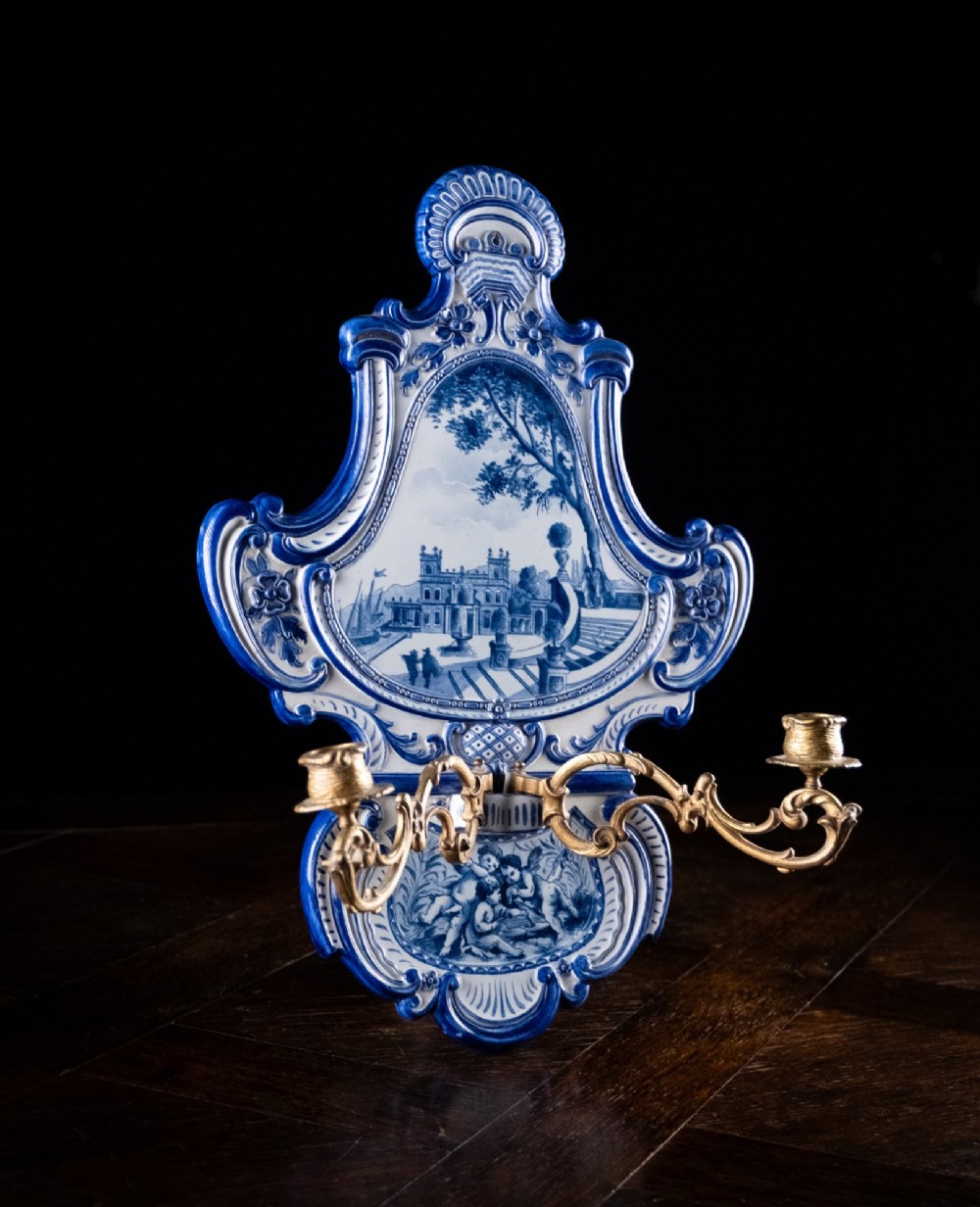 A Rococo Revival blue and white glazed ceramic wall plaque depicting an architectural scene and cherubs, fitted with two gilt ormolu candle branches. View XI