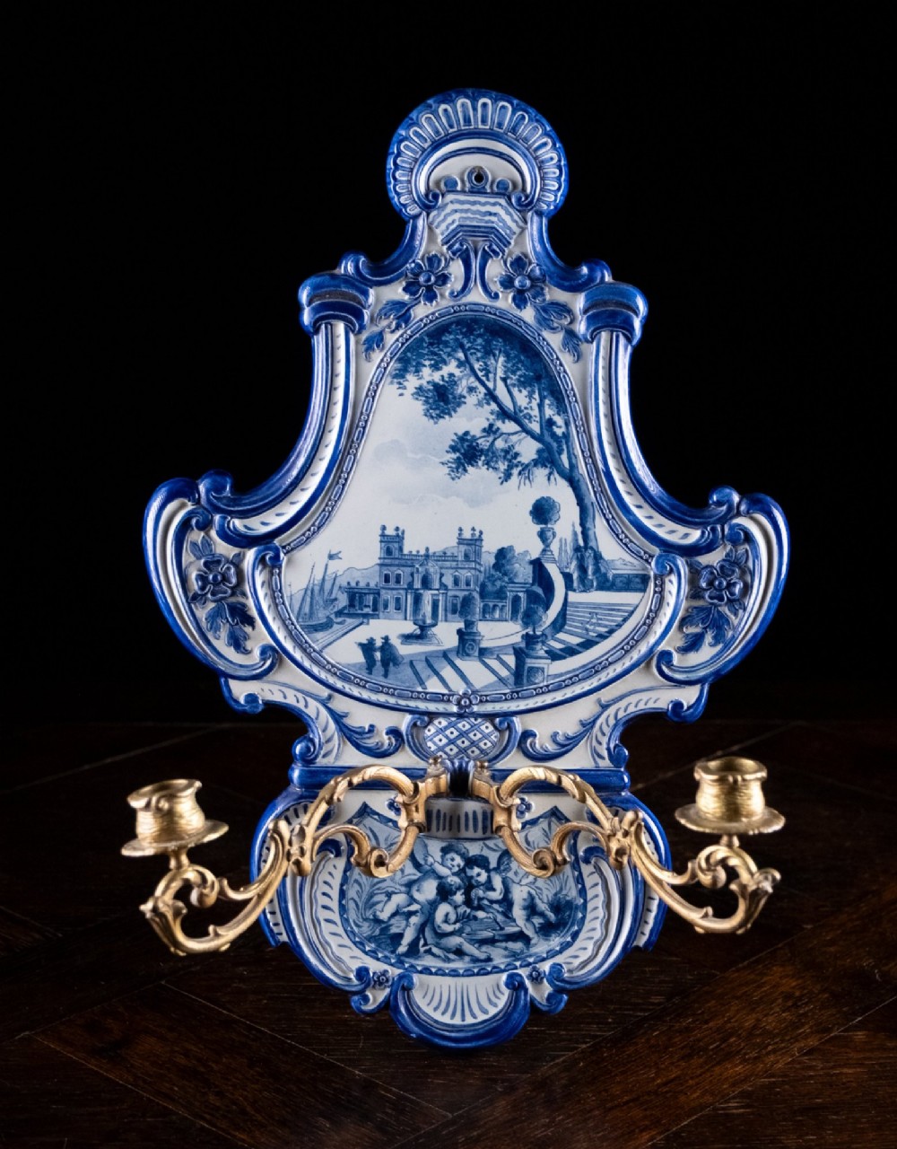 A Rococo Revival blue and white glazed ceramic wall plaque depicting an architectural scene and cherubs, fitted with two gilt ormolu candle branches. View XII