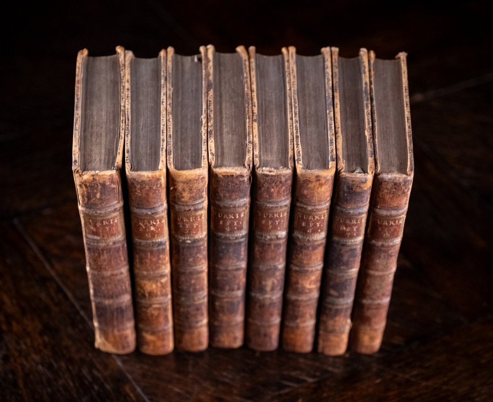 An eight-volume set of 18th-century leather-bound books standing upright with visible raised spine bands and gilt titles. View IV