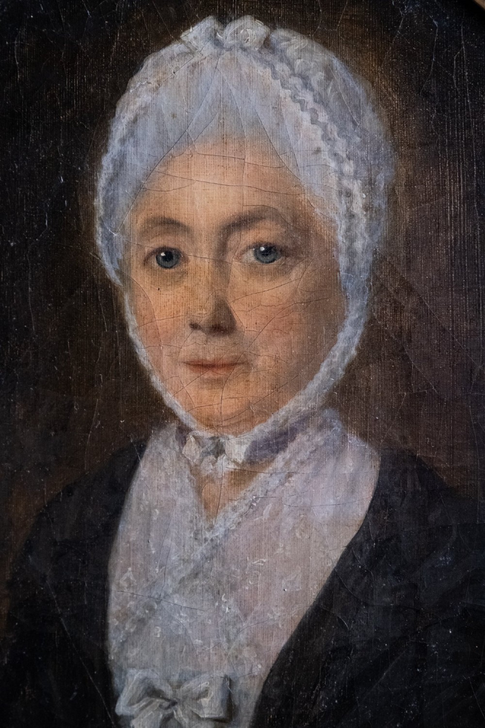 An 18th-century oil painting depicting a lady in a white lace cap and dark garment, framed within a gilded oval slip set inside a rectangular dark wooden frame. View II