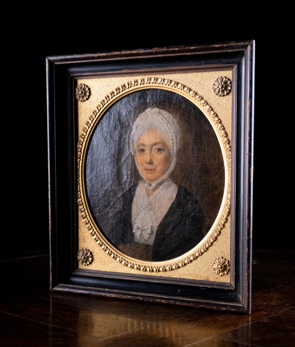 An 18th-century oil painting depicting a lady in a white lace cap and dark garment, framed within a gilded oval slip set inside a rectangular dark wooden frame. View VI