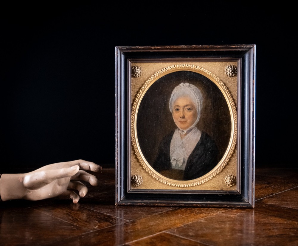 An 18th-century oil painting depicting a lady in a white lace cap and dark garment, framed within a gilded oval slip set inside a rectangular dark wooden frame. View VII