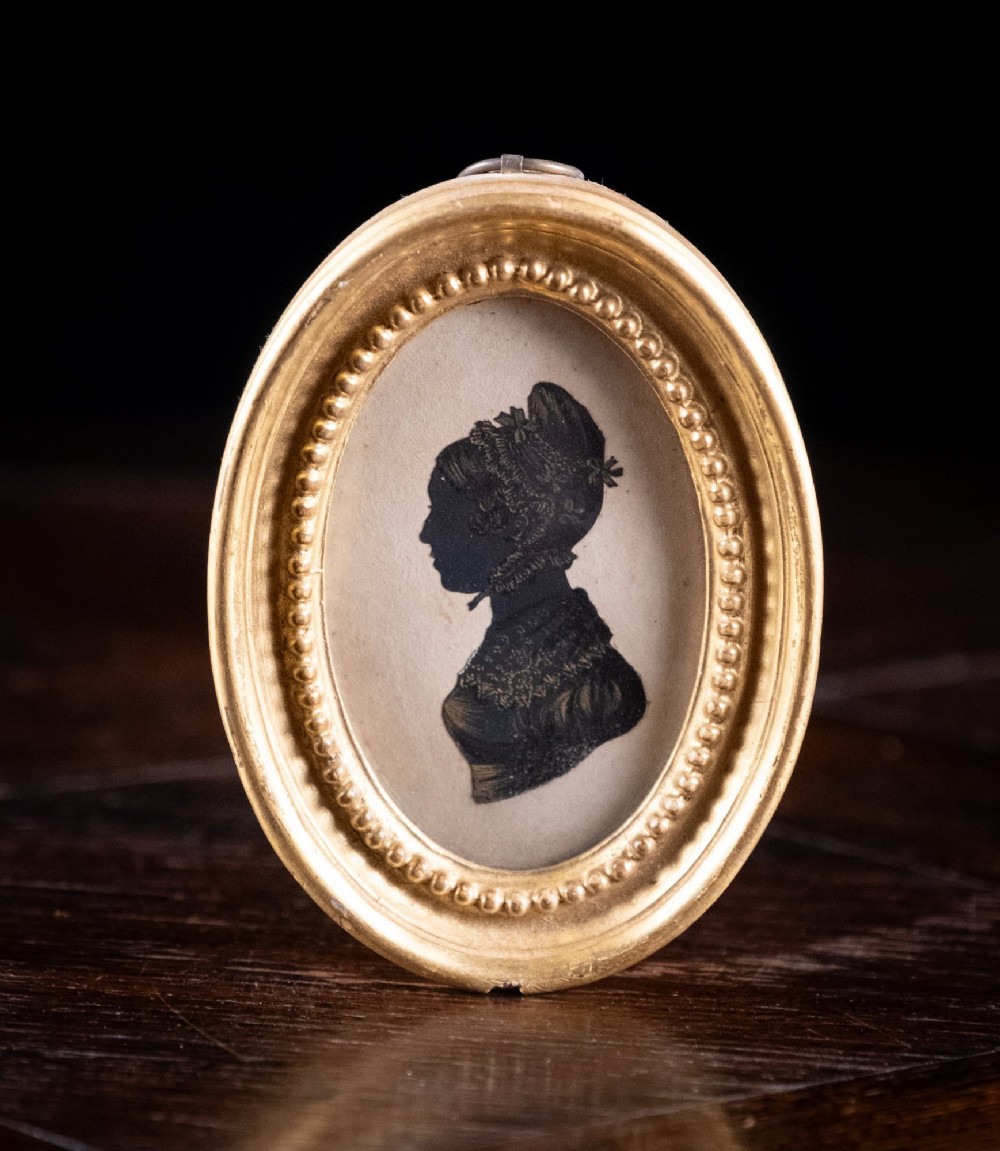 An oval framed Regency-era miniature silhouette depicting the profile of a lady embellished with fine gold detailing, set within a beaded gilt frame.