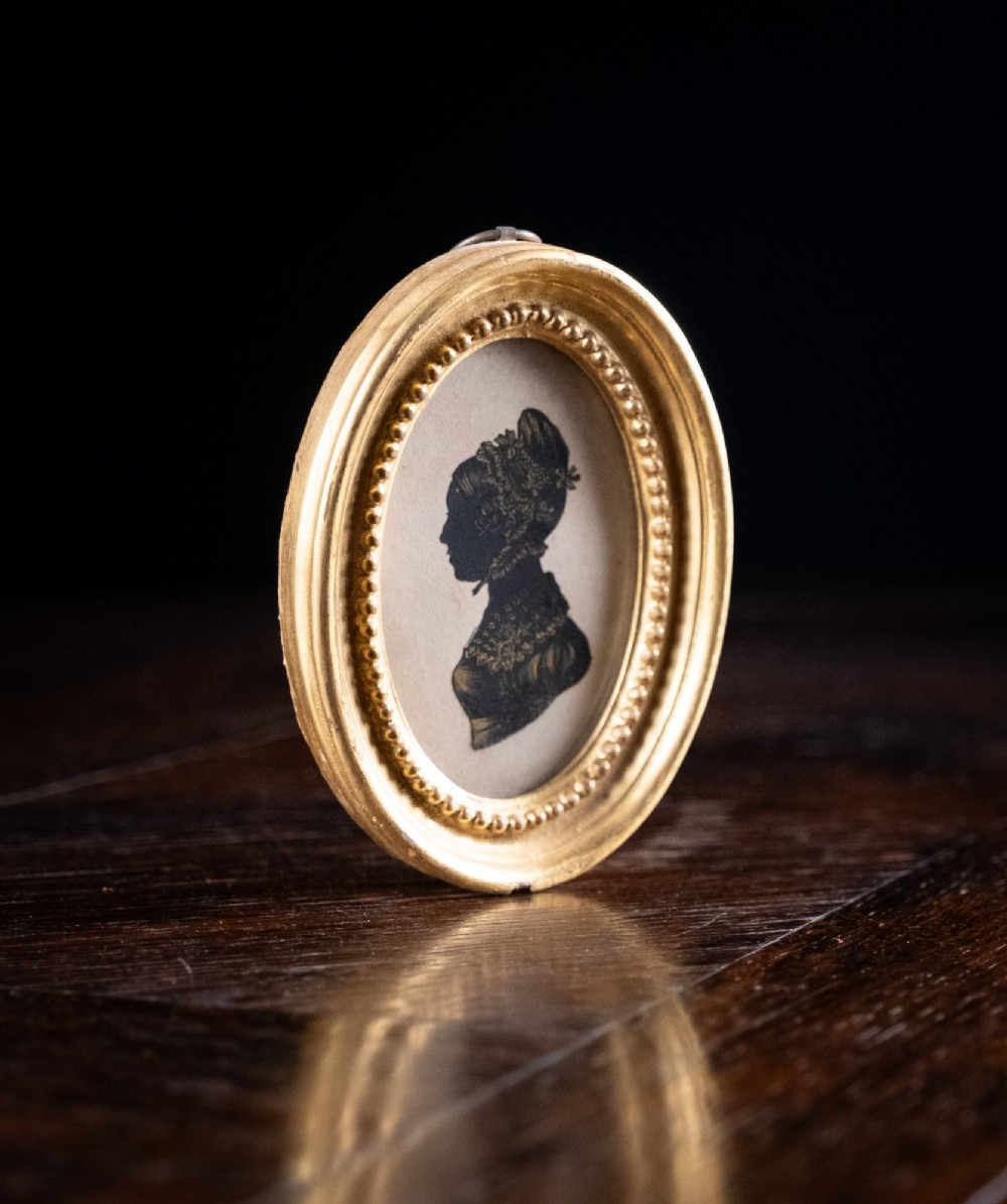 An oval framed Regency-era miniature silhouette depicting the profile of a lady embellished with fine gold detailing, set within a beaded gilt frame. View III
