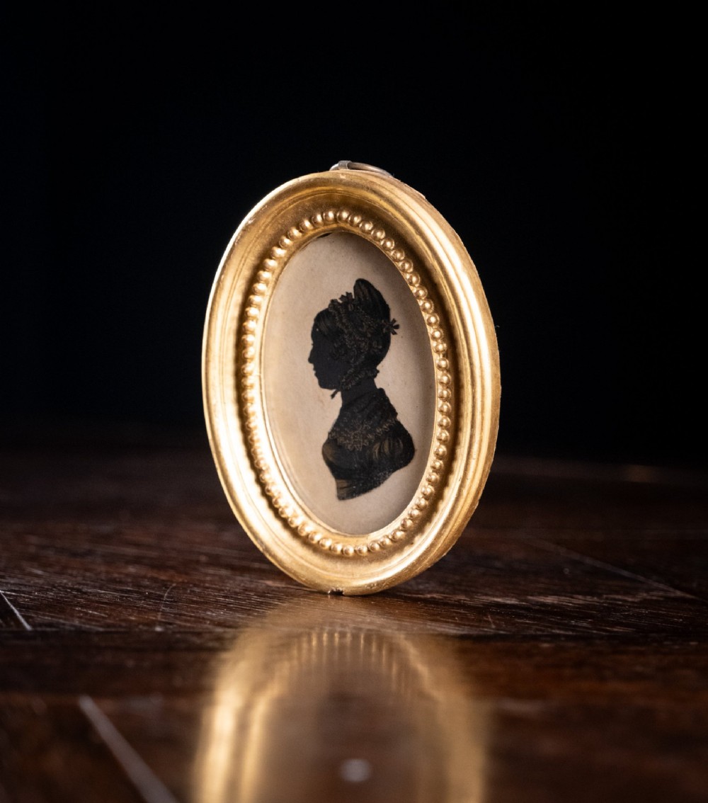An oval framed Regency-era miniature silhouette depicting the profile of a lady embellished with fine gold detailing, set within a beaded gilt frame. View IV