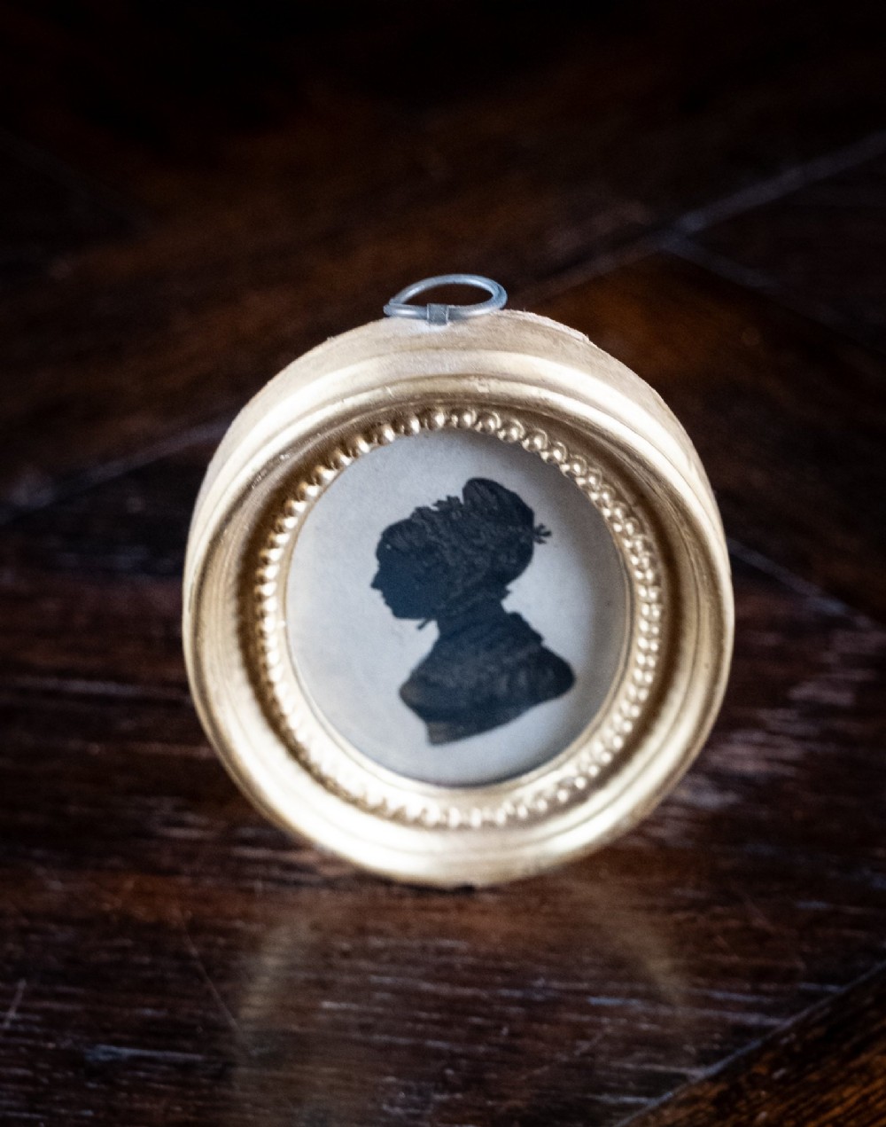 An oval framed Regency-era miniature silhouette depicting the profile of a lady embellished with fine gold detailing, set within a beaded gilt frame. View V